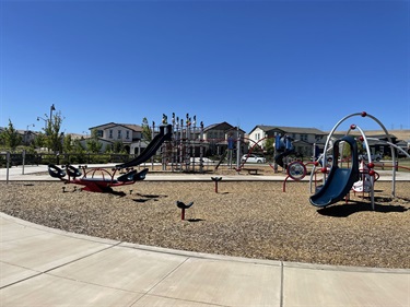 Playground with slides, climbing structures, spinning play features, and interactive equipment on a wood chip surface, bordered by a concrete walkway and surrounded by open grassy areas in a community park.