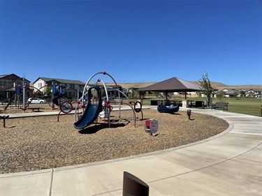Playground with a curved slide, climbing structures, interactive play features, and spring riders on a wood chip surface, with a covered picnic shelter, walking paths, and open grassy areas in a community park.