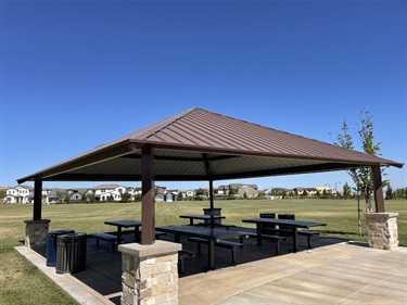 Large covered picnic pavilion with multiple picnic tables on a concrete pad, surrounded by open grassy areas, walking paths, and young trees in a community park under a clear blue sky.