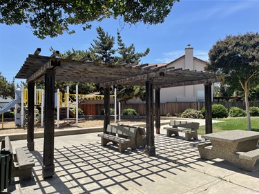 Pergola-covered picnic area with concrete picnic tables on a paved patio, adjacent to a playground and landscaped park setting with mature trees.