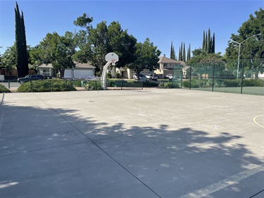 Outdoor basketball court with a concrete playing surface and a single basketball hoop, bordered by mature shade trees, chain-link fencing, landscaped planting beds, and open park space under a clear blue sky.