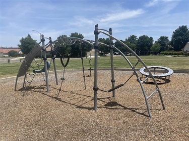 Modern playground climbing structure with arched overhead climbing frames, rope climbing elements, hanging rings, balance features, and a rotating play spinner on a wood chip safety surface, surrounded by open grassy areas in a community park.