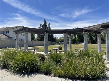 Pergola-covered seating area with picnic tables, landscaped shrubs, and an open grassy lawn under a blue sky.