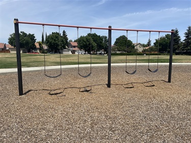 Row of swing sets with rubber seats on a mulch surface, with houses and trees in the background.