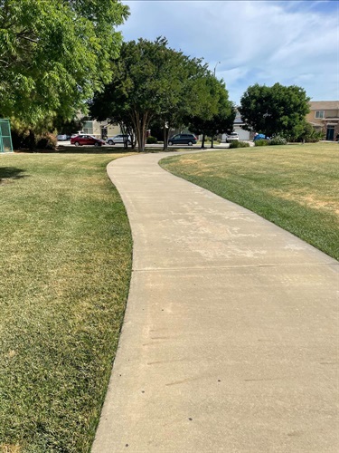 Concrete walking path winding through a community park, bordered by open grassy areas, mature shade trees, and landscaped park space, with nearby residential buildings visible in the background under a partly cloudy sky.