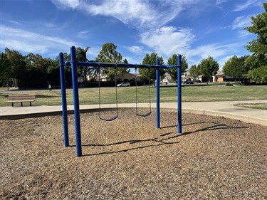 Two-bay swing set with belt swings on a wood chip safety surface, surrounded by open grassy areas, concrete walking paths, park benches, and mature shade trees in a community park under a partly cloudy sky.
