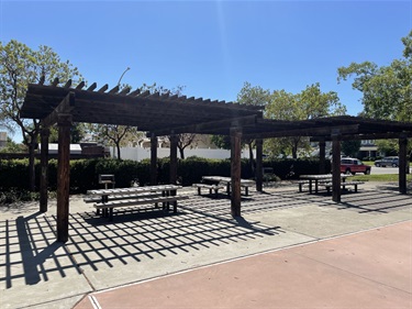 Wooden pergola shade structure with picnic tables underneath, set on a paved patio in a park with a red car and trees in the background.