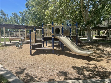 Playground with slides, climbing features, and activity panels on a wood chip surface, shaded by mature trees with picnic shelters nearby in a community park.