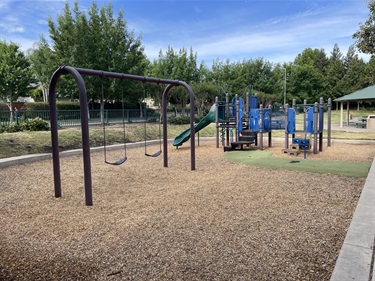 Playground with a multi-level climbing structure, slides, and a two-bay swing set with belt swings on a wood chip safety surface, bordered by a concrete walkway and surrounded by mature trees, open grassy areas, and a covered picnic pavilion in a community park.