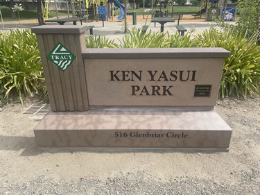 A stone and wood monument sign for Ken Yasui Park, with a small logo at the top and the park's street address printed below the name.