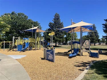 A playground with blue and yellow shade canopies over play structures, slides, and swings, set on a wood-chip surface with trees in the background.
