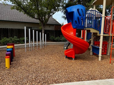 Colorful playground structure with a red spiral slide and blue climbing tower, on a mulch surface with a building and trees in the background.