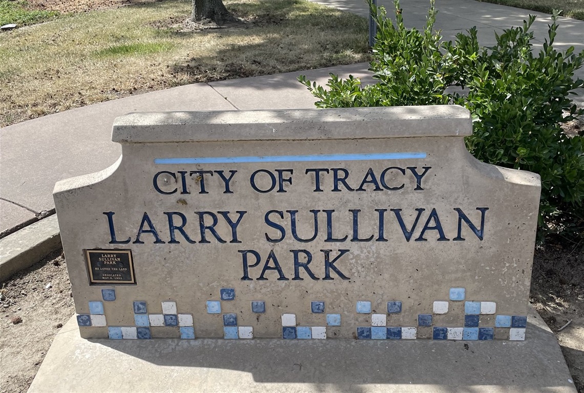 Stone entrance sign reading 'City of Tracy, Larry Sullivan Park' with a blue tile mosaic border, framed by greenery.