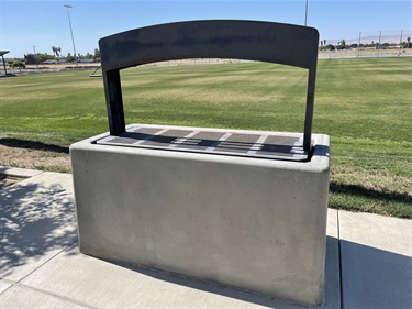Concrete park trash and recycling receptacle with separate disposal openings, positioned beside a concrete walkway overlooking open grassy fields in a community park.