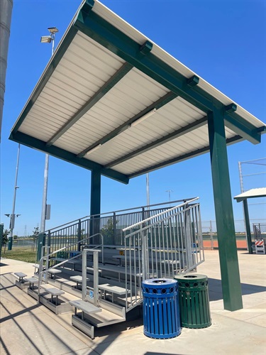 Covered spectator bleachers with metal seating beneath a shade canopy, overlooking baseball fields, with trash and recycling bins nearby in a community sports park.