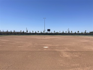 Baseball field with a dirt infield, pitcher's mound, and open outfield, viewed from behind home plate with a scoreboard visible in the distance under a clear blue sky.