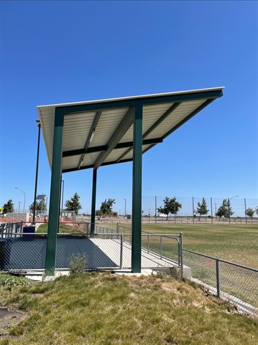 Covered spectator viewing area with a metal shade canopy beside a chain-link fence, overlooking open sports fields in a community park under a clear blue sky.