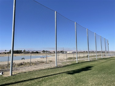 Tall ball stop netting supported by metal poles along the edge of a sports field, with open grassy areas and a community sports complex visible under a clear blue sky.