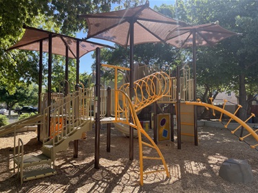 Playground with climbing structures, overhead climbing arches, activity panels, elevated platforms, and shade canopies on a wood fiber safety surface, surrounded by mature trees