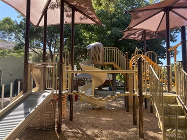 Playground with elevated platforms, climbing structures, a spiral slide, bridges, and shade canopies on a wood fiber safety surface, surrounded by mature trees.