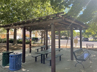 Pergola-covered picnic area with multiple picnic tables and benches on a paved patio, surrounded by mature trees, landscaped park grounds, and nearby parking.