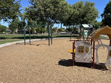 Nature-themed playground with playhouses, activity panels, and a two-bay swing set on a wood chip surface, surrounded by mature shade trees, open grassy areas, and walking paths in a community park.
