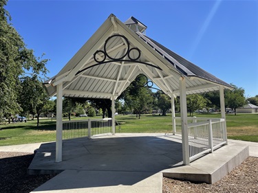 Decorative covered gazebo with open seating space, white railings, and ornamental metal detailing, surrounded by open grassy lawns, mature shade trees, and walking paths in a community park.