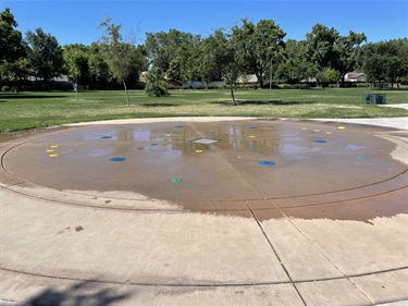 Circular splash pad with ground-level water spray features on a concrete surface, surrounded by open grassy lawns, mature trees, and walking paths in a community park.