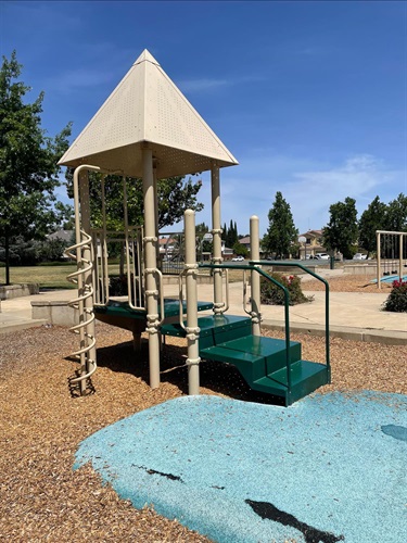 Small playground structure with a peaked tan roof, teal steps and platform, and a rope climbing net, on a mulch and rubber surface with a grassy field in the background.