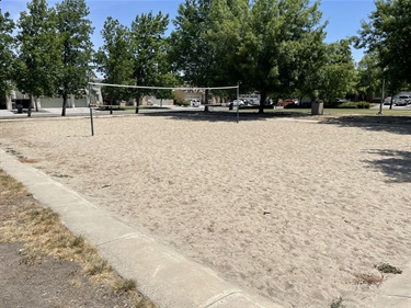 Sand volleyball court with a net, bordered by a paved path, with a shade shelter and trees in the background.
