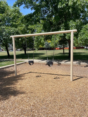 Wooden swing set with two rubber seats over a mulch surface, with a lawn and trees in the background.