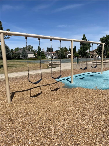 Swing set with rubber seats and a glider swing over a mulch surface, with a teal rubber play area and houses in the background.