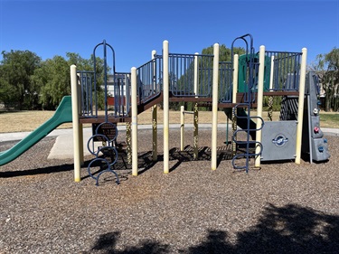 Playground climbing structure with a green slide, spring rockers, and a tower, on a mulch surface with a grassy field and trees in the background.
