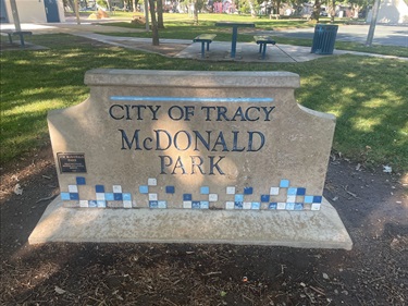 Stone sign reading ‘City of Tracy McDonald Park’ with decorative blue and white tiles along the base, surrounded by grass and trees in a park setting.