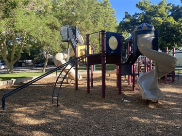 Playground with climbing structures, slides, and elevated play platforms surrounded by wood-chip surfacing and mature shade trees.