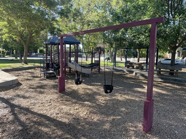 Swing set with multiple belt swings adjacent to a playground, surrounded by wood-chip surfacing and shaded by mature trees.