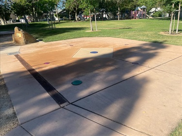 Splash pad with ground-level water spray features set into a concrete play surface, surrounded by open grassy areas and shade trees in a park setting.
