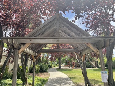 Wooden gazebo structure with a peaked roof, surrounded by a red-leafed tree and a paved path leading through a garden.