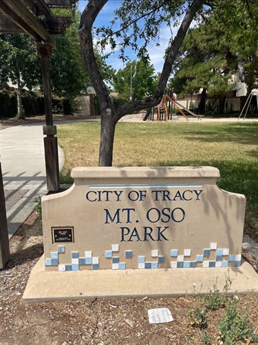 Stone entrance sign reading 'City of Tracy, Mt. Oso Park' with a blue tile mosaic border, with a playground and trees in the background.