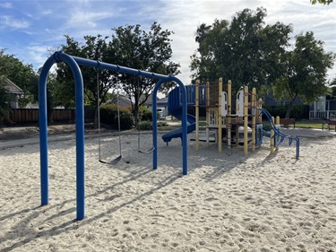 Blue swing set and playground climbing structure on a sand surface, with houses and trees in the background.