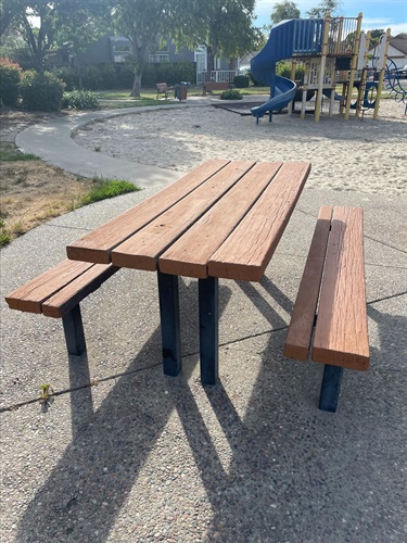 Wooden picnic table with attached benches on a paved patio, with a playground slide and trash can visible in the background.