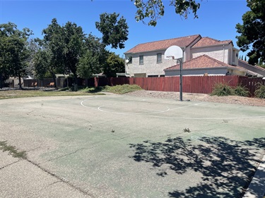 Empty basketball court with a hoop, bordered by a wooden fence, trees, and a two-story house in the background.