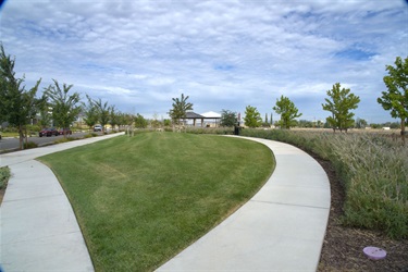 Curving concrete walking paths surrounding a landscaped grassy lawn, with young trees, ornamental plantings, and open natural scenery under a partly cloudy sky.