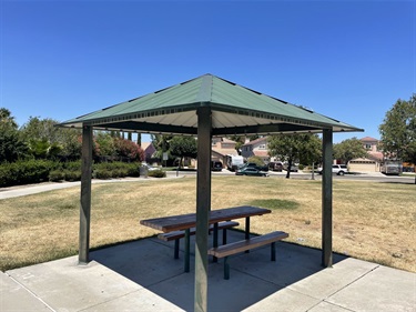 Green-roofed gazebo shelter with a picnic table on a paved patio, set on a lawn with houses in the background.