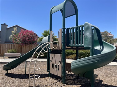 Green playground structure with two slides, a climbing wall, and an activity panel, on a mulch surface with a fence and houses in the background.