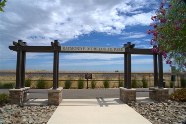 Decorative park overlook with a wooden pergola, benches, paved walkway, and ornamental landscaping, offering views of an open water feature under a cloudy sky.