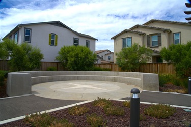 Circular concrete gathering area with low seating walls, surrounded by landscaped planting beds and neighboring residential homes.