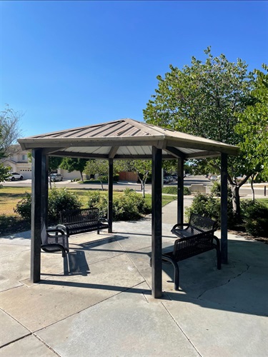 Covered gazebo with built-in benches on a paved patio, surrounded by landscaped planting beds, mature trees, and park walkways.