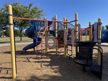 Playground with climbing structures, activity panels, overhead play elements, and spiral slides on a wood fiber safety surface, surrounded by open park space.