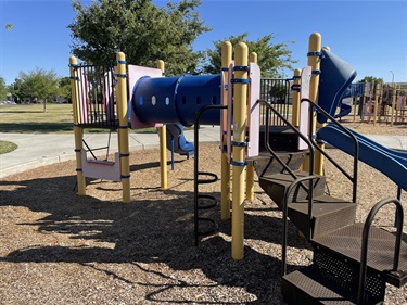 Playground with elevated platforms, climbing structures, a blue crawl tunnel, slides, and accessible ramps on a wood fiber safety surface, surrounded by open park space.